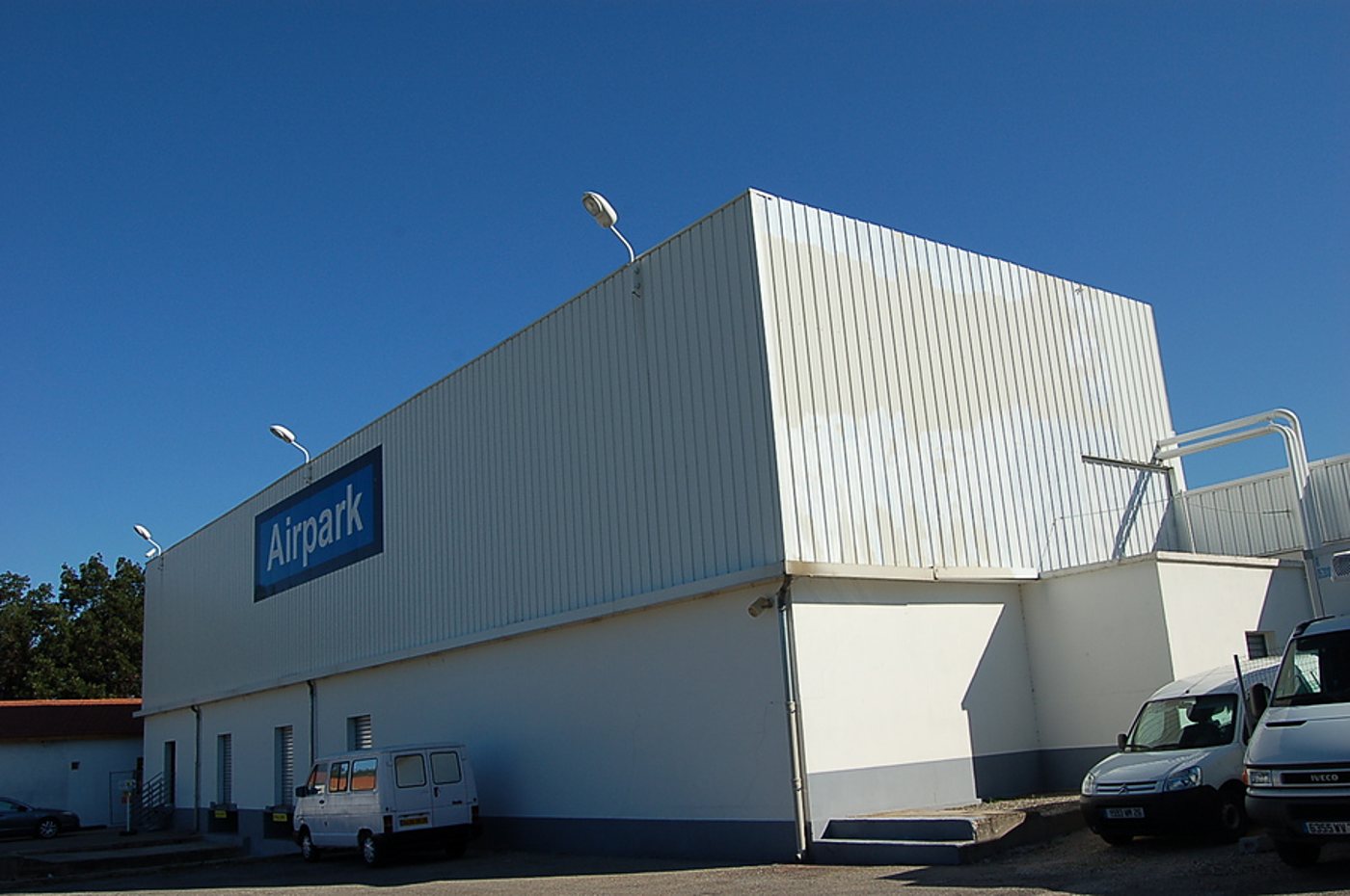 Façade d'un grand entrepôt blanc portant une enseigne bleue Airpark, avec des camionnettes blanches garées sur le parking sous un ciel bleu.