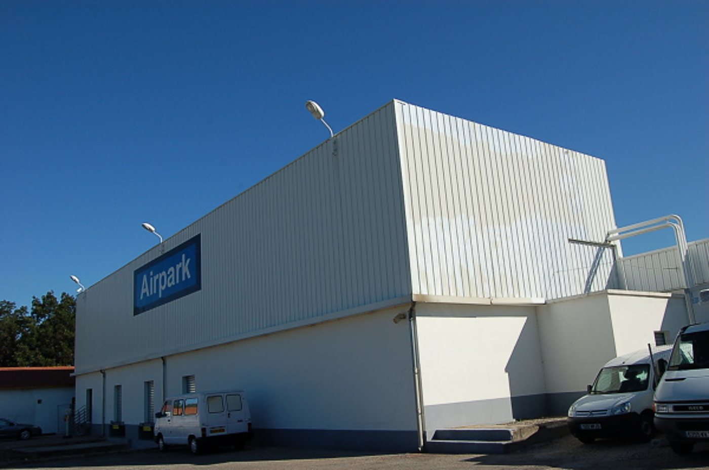Un grand bâtiment industriel blanc à bardage métallique porte une enseigne bleue Airpark, avec des camionnettes garées devant sous un ciel bleu.