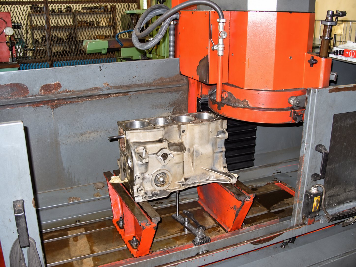Un bloc moteur en aluminium est posé sur des supports rouges sous la tête d'une grande rectifieuse industrielle dans un atelier de mécanique.
