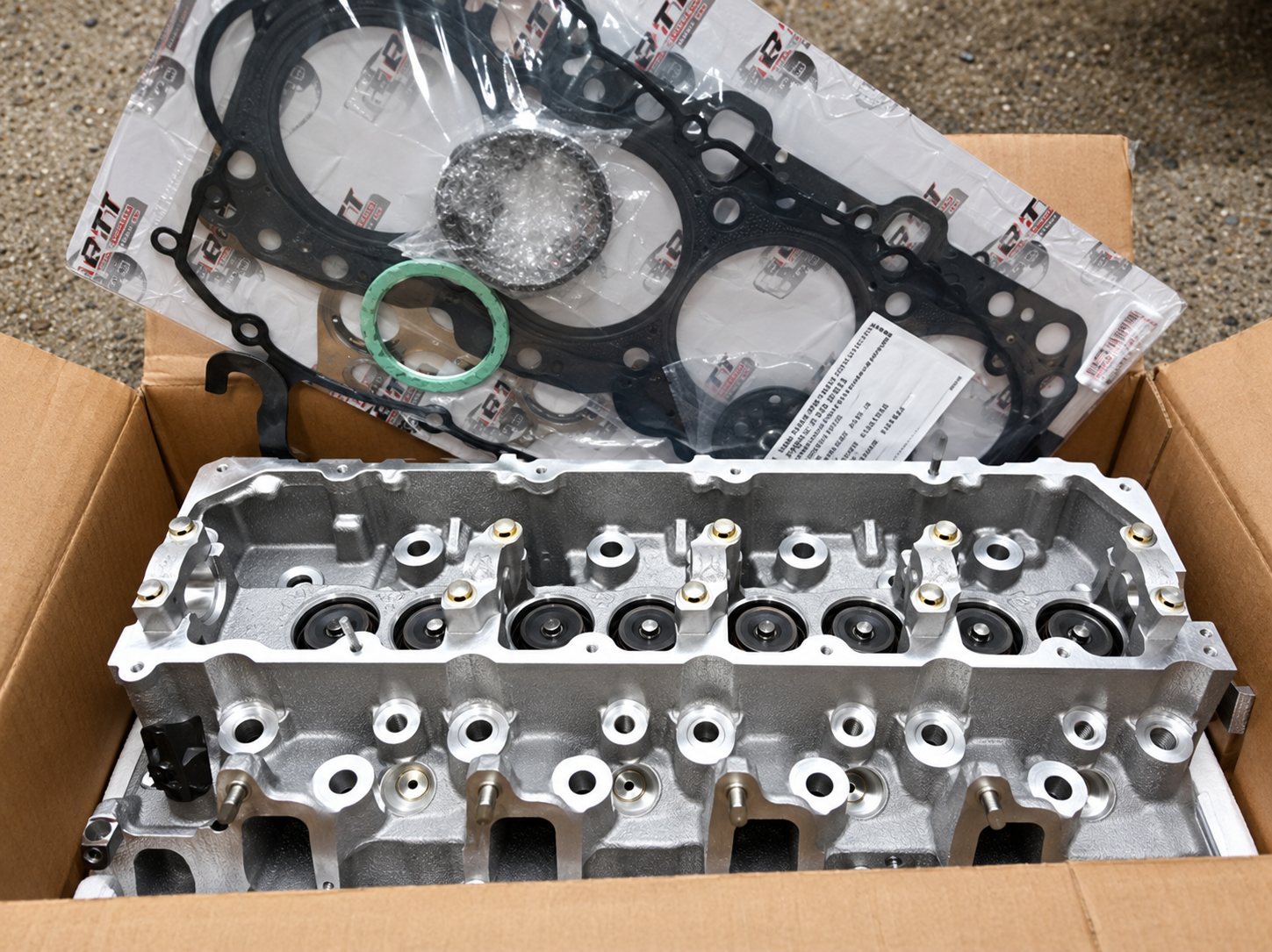 Une culasse de moteur en aluminium posée dans un carton ouvert, accompagnée d'un joint de culasse et de joints d'étanchéité.