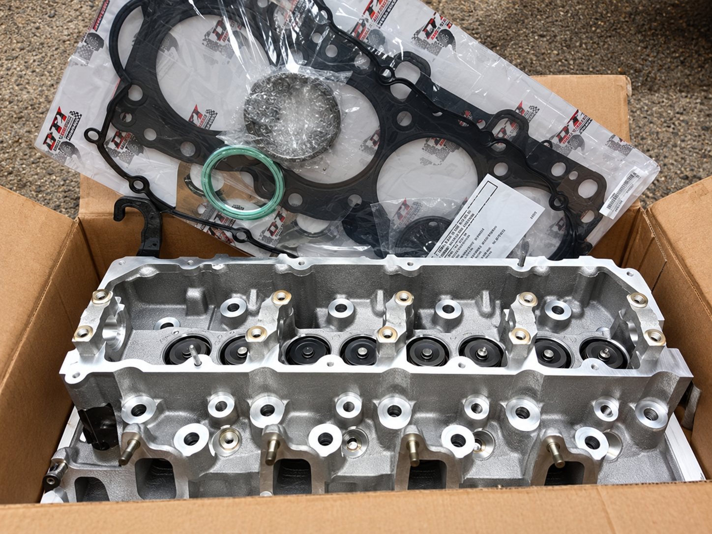 Une culasse de moteur en aluminium posée dans un carton ouvert, accompagnée d'un joint de culasse et d'autres joints au-dessus.