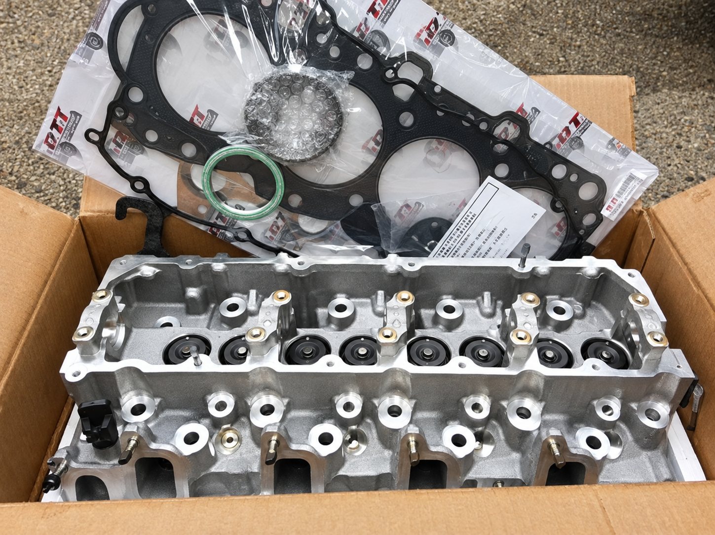 Une culasse de moteur en aluminium posée dans un carton ouvert, accompagnée d'un joint de culasse et de petits joints d'étanchéité.