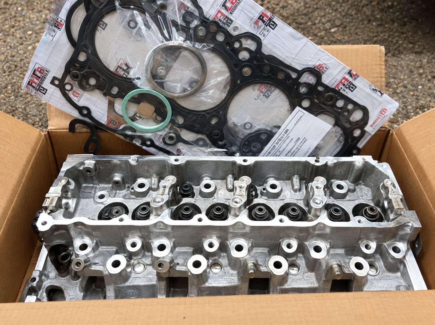 Une culasse de moteur en aluminium posée dans un carton ouvert, avec un jeu de joints visible derrière elle.