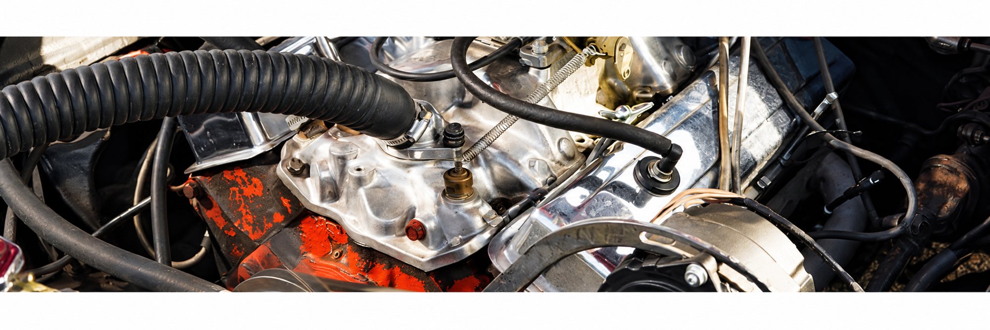 Vue rapprochée d'un moteur V8 ancien avec carburateur et pièces chromées, entouré de durites noires et de câbles, bloc moteur peint en orange.