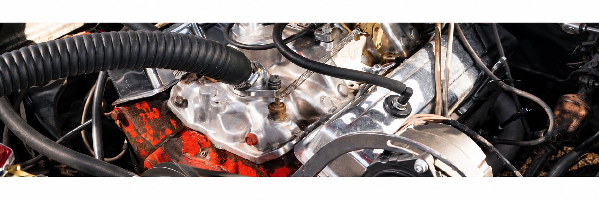 Gros plan sur le compartiment moteur d'un véhicule ancien, avec un carburateur chromé, des durites noires et un bloc moteur peint en orange.