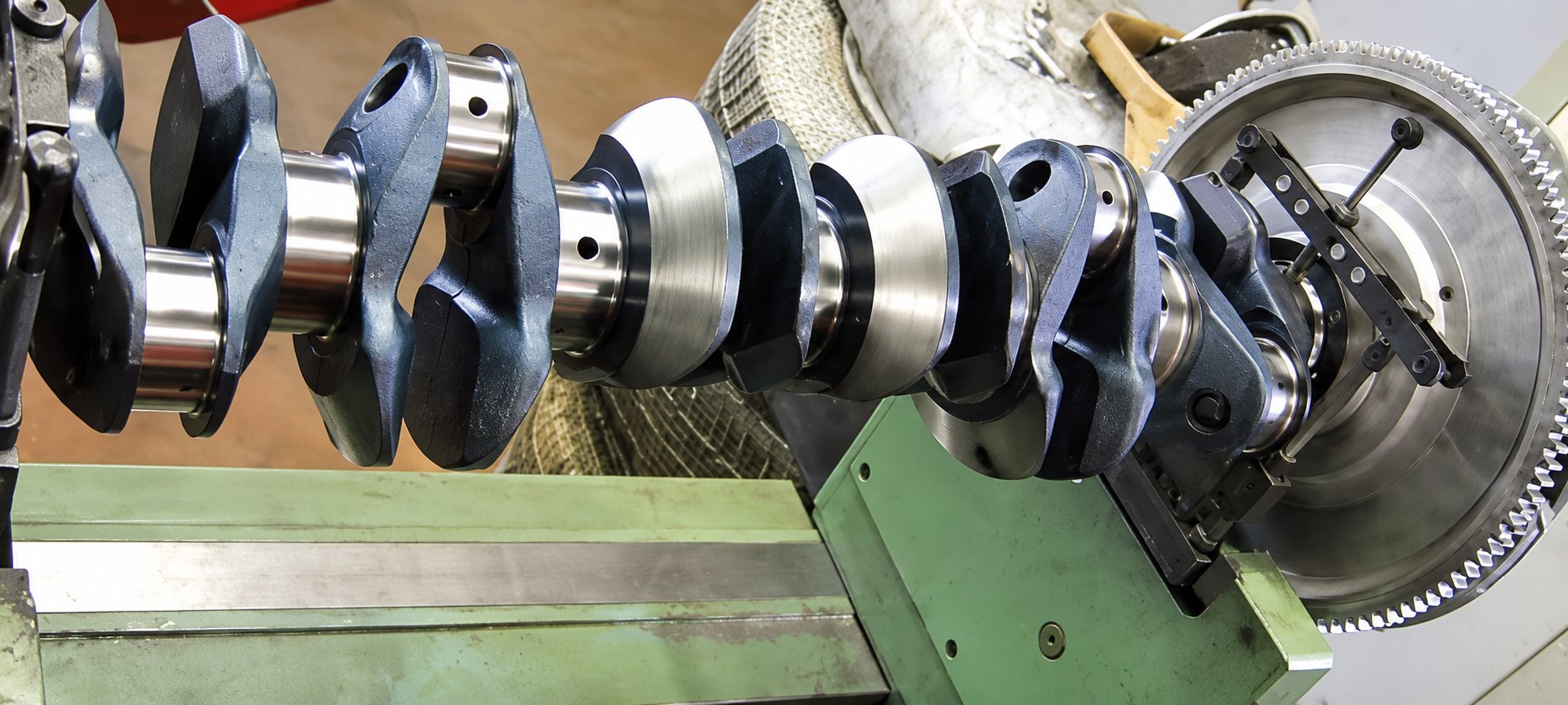 Un vilebrequin en acier est installé sur une machine d'atelier verte, avec un volant moteur denté à droite.